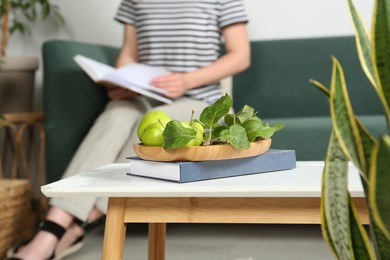 Woman reading book in living room, focus on table with apples