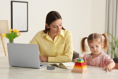 Photo of Woman working remotely. Mother with laptop and daughter at desk