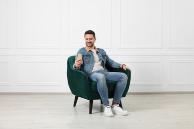 Happy man sitting in armchair and using smartphone indoors