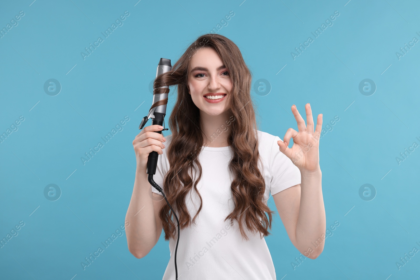 Photo of Beautiful woman showing OK gesture while using curling hair iron on light blue background
