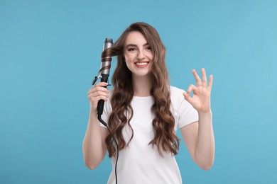 Photo of Beautiful woman showing OK gesture while using curling hair iron on light blue background