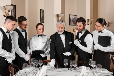 Photo of Senior man wearing formal suit teaching trainees in restaurant. Professional butler courses