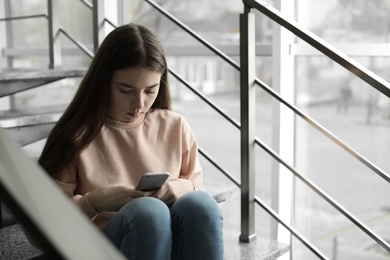 Photo of Upset teenage girl with smartphone sitting on stairs indoors. Space for text