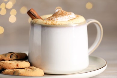Photo of Cup of delicious eggnog with cinnamon and cookies on table against festive lights, closeup