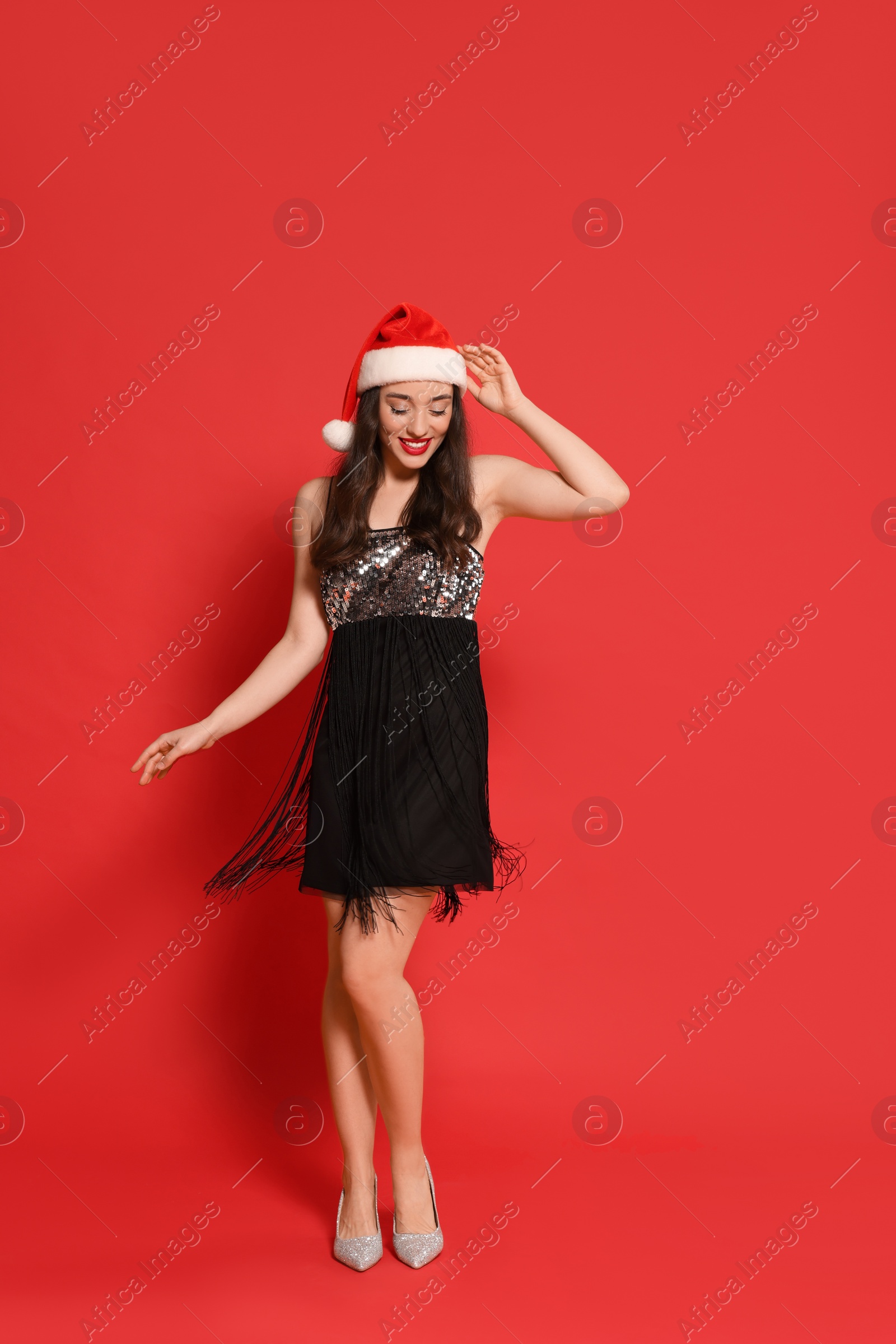 Photo of Christmas celebration. Beautiful young woman in stylish dress and Santa hat on red background