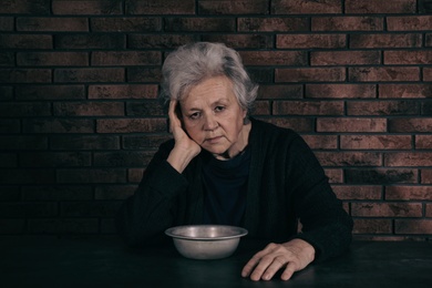 Poor mature woman with bowl sitting at table