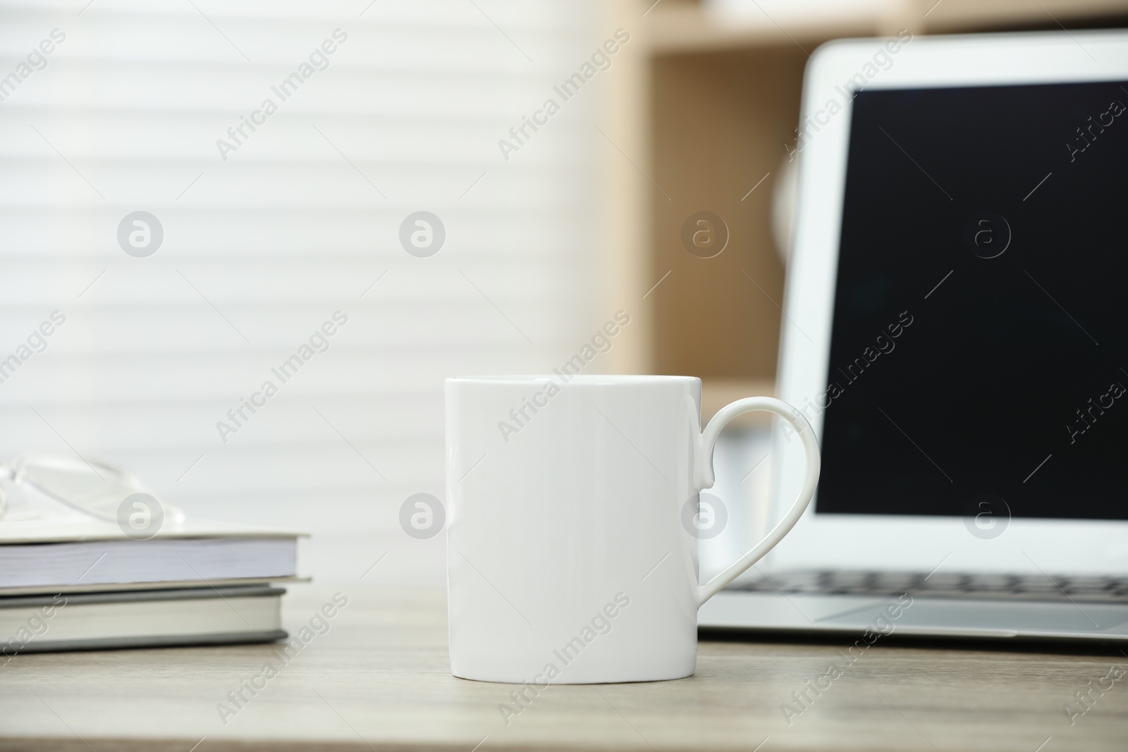 Photo of White ceramic mug, notebooks and laptop on wooden table indoors