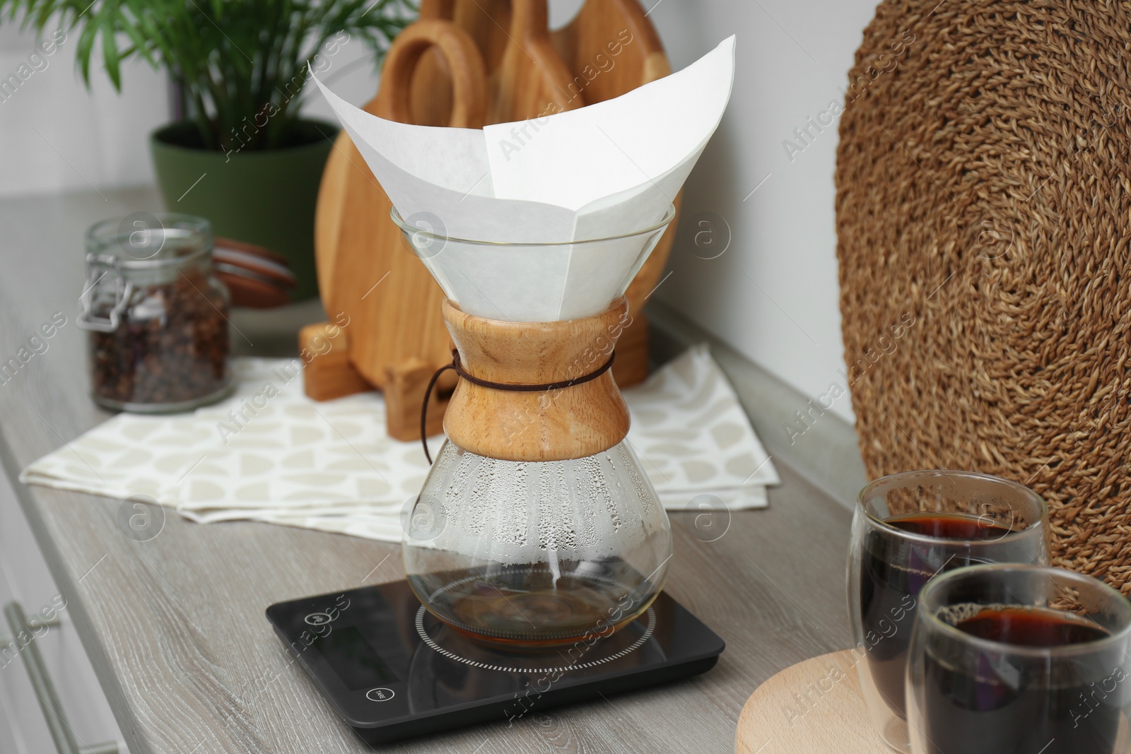 Photo of Glass chemex coffeemaker with paper filter, scales and glasses of coffee on wooden countertop