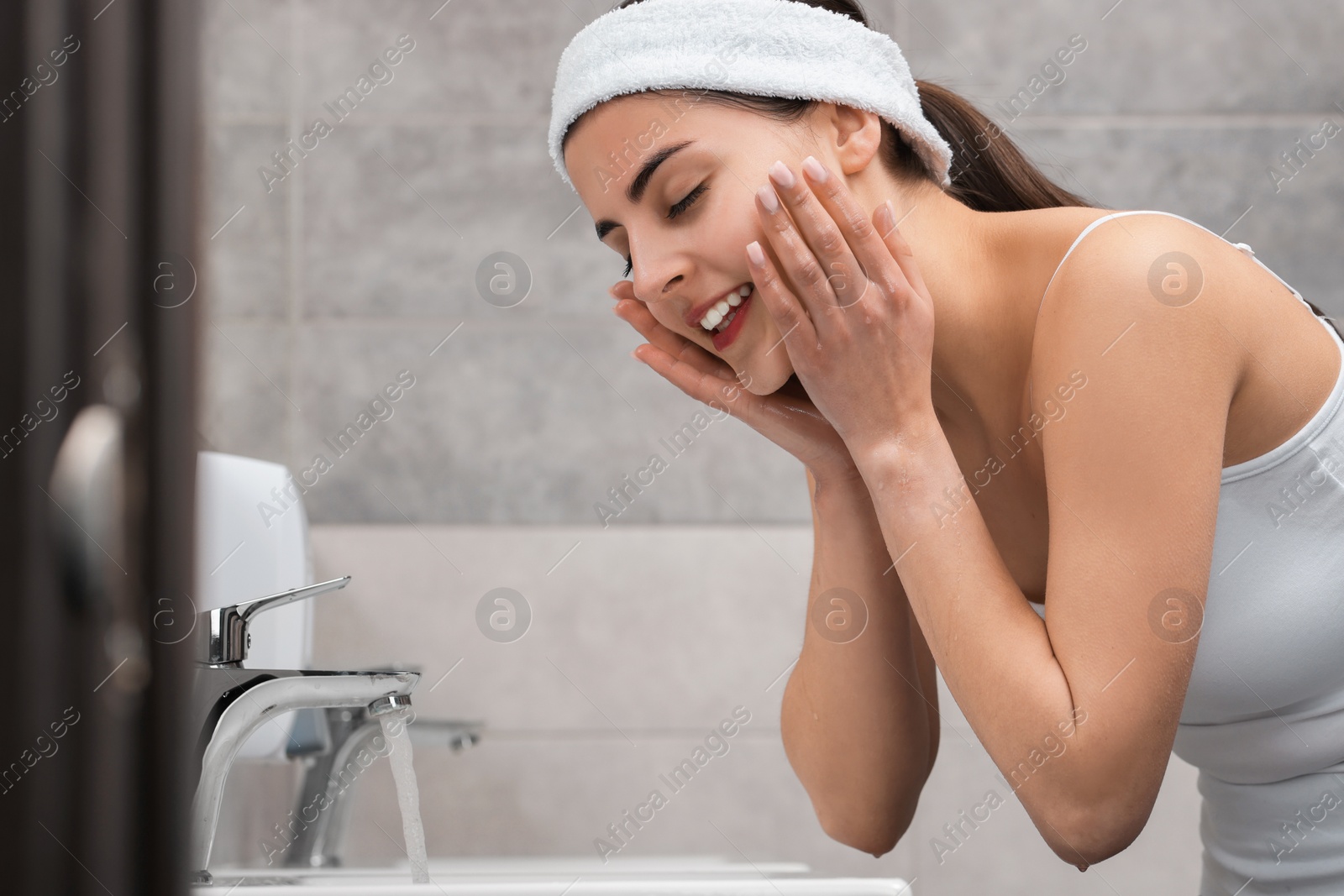Photo of Young woman with headband washing her face in bathroom