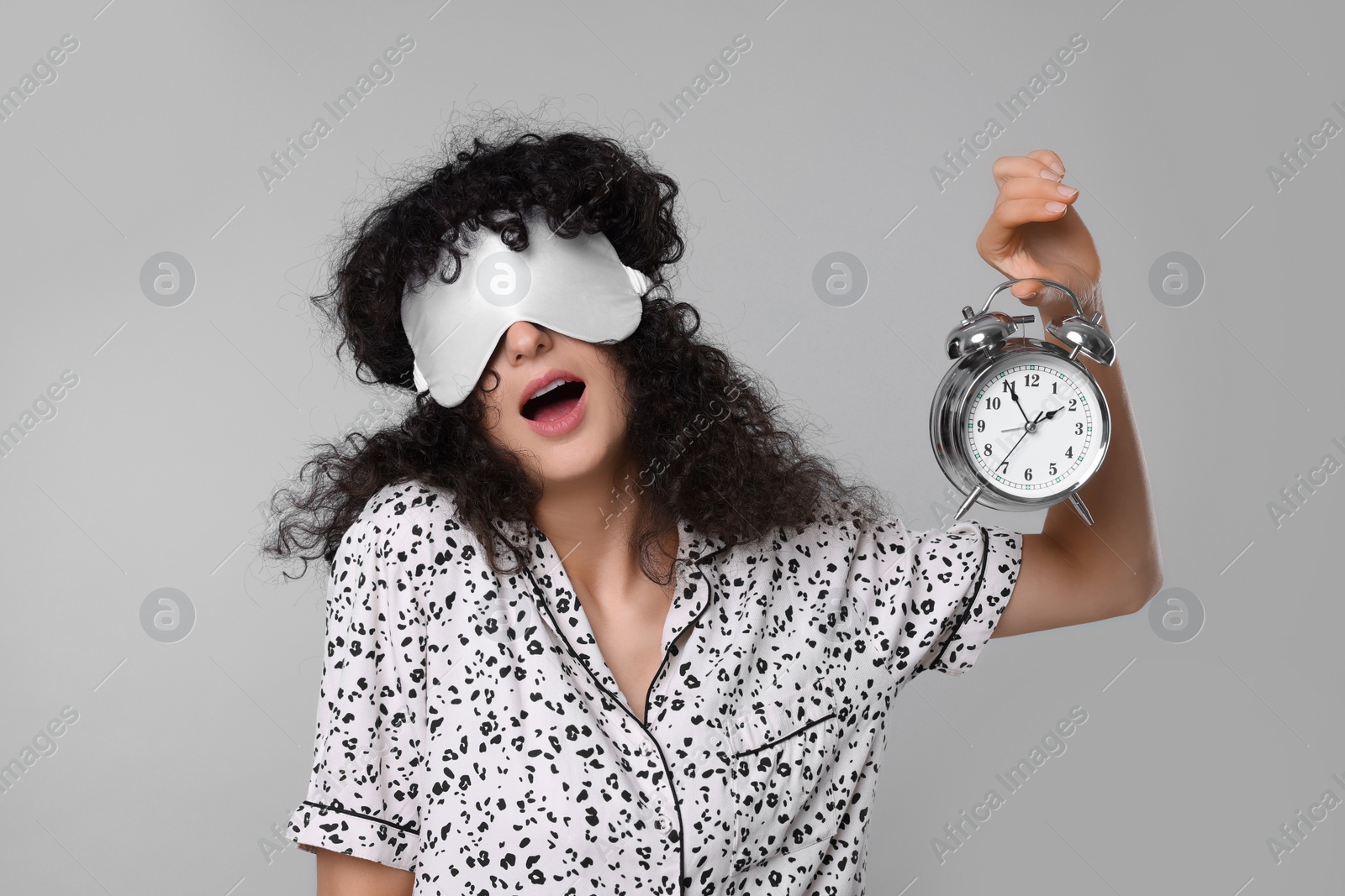 Photo of Tired young woman with sleep mask and alarm clock yawning on light grey background. Insomnia problem