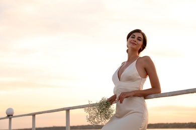 Gorgeous bride in beautiful wedding dress with bouquet near river on sunset. Space for text