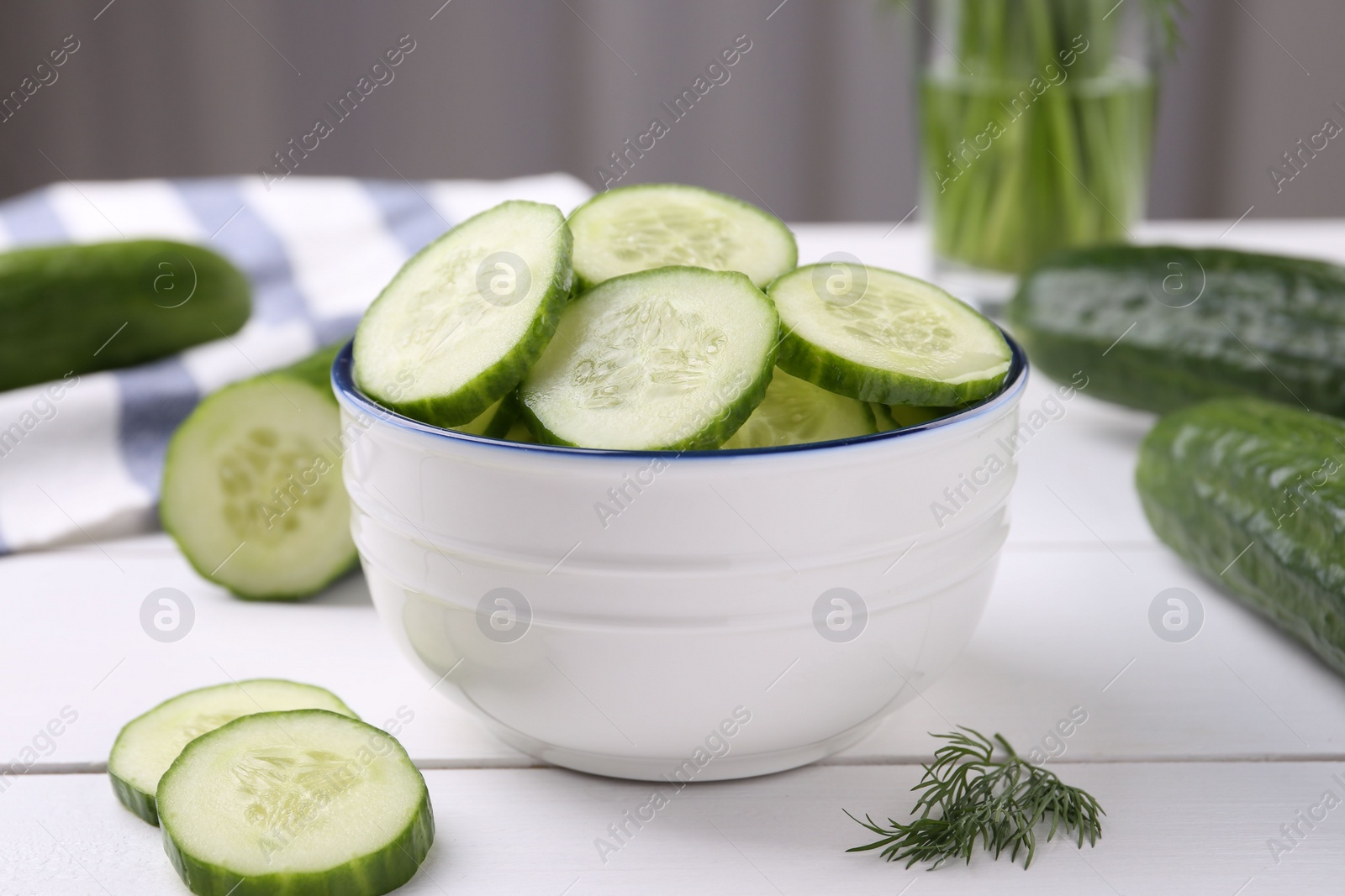 Photo of Cut cucumber in bowl, fresh vegetables and dill on white wooden table, closeup