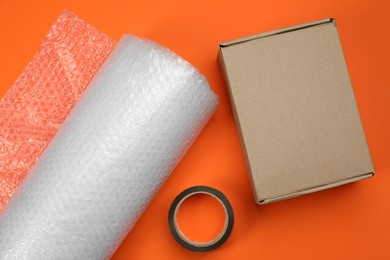 Bubble wrap roll, cardboard box and tape on orange background, flat lay