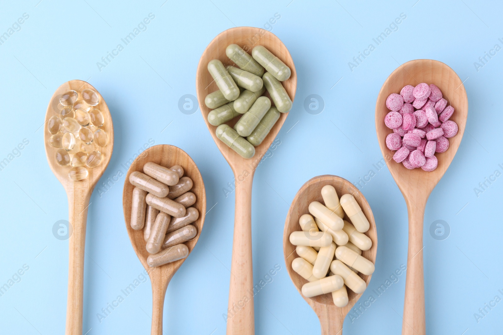 Photo of Different vitamin pills in wooden spoons on light blue background, flat lay