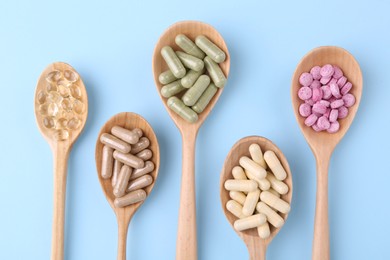 Different vitamin pills in wooden spoons on light blue background, flat lay