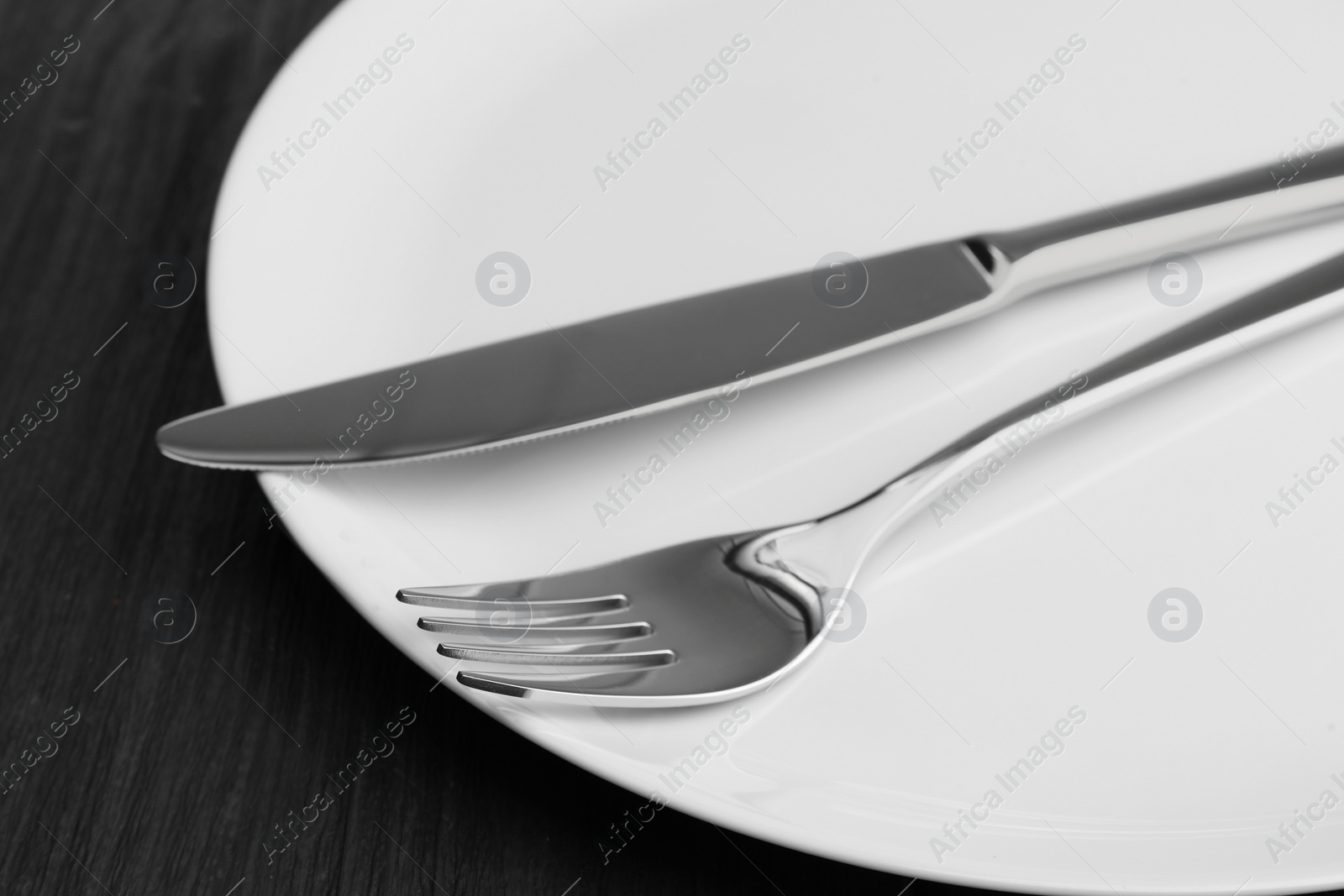 Photo of Clean plate, fork and knife on black wooden table, closeup