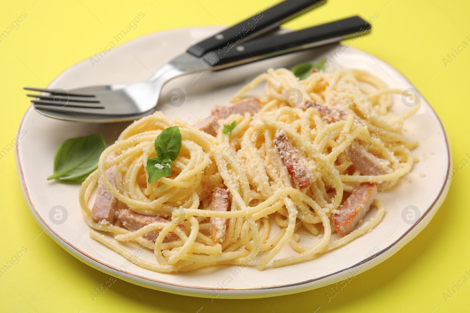 Photo of Plate of tasty pasta Carbonara with basil leaves, fork and spoon on yellow background, closeup