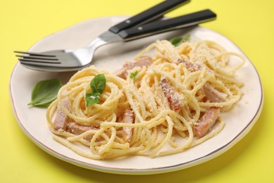 Photo of Plate of tasty pasta Carbonara with basil leaves, fork and spoon on yellow background, closeup