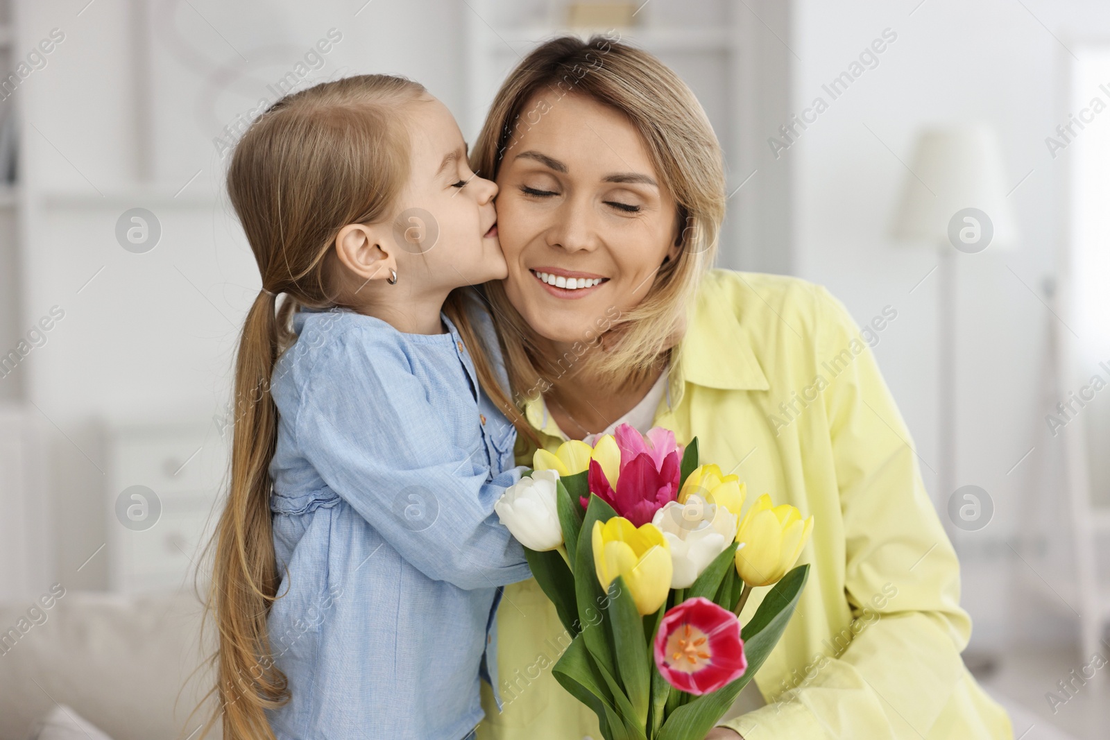 Photo of Little daughter kissing and congratulating her mom with Mother`s Day at home. Woman holding bouquet of beautiful tulips