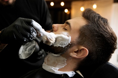 Professional hairdresser applying shaving foam onto client's skin in barbershop