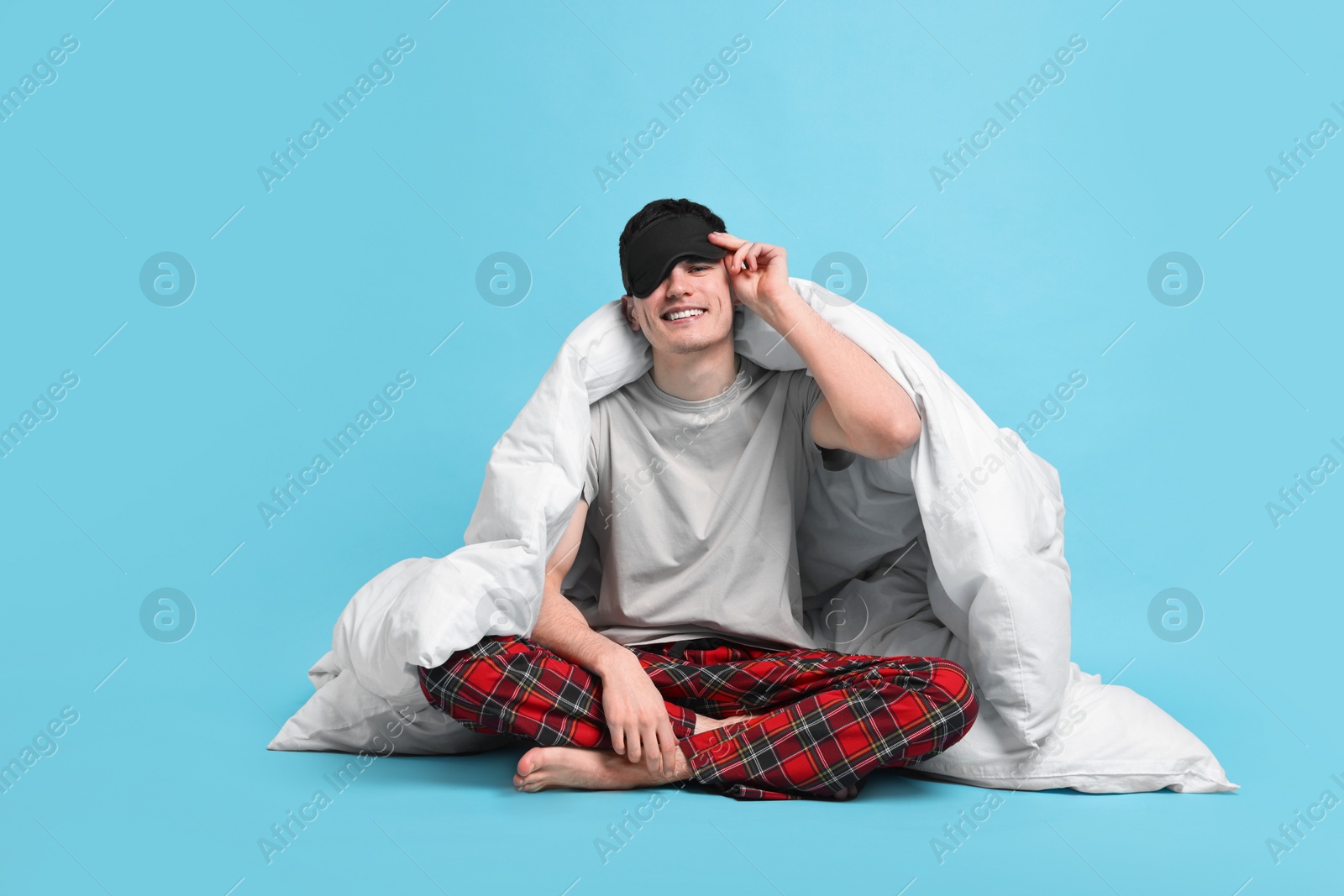 Photo of Happy man in pyjama and sleep mask wrapped in blanket on light blue background