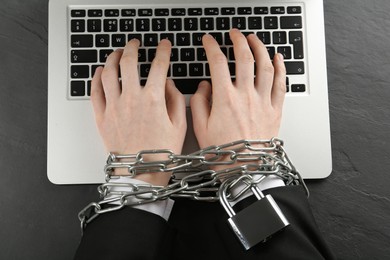 Photo of Internet addiction. Man typing on laptop with chained hands at black table, top view