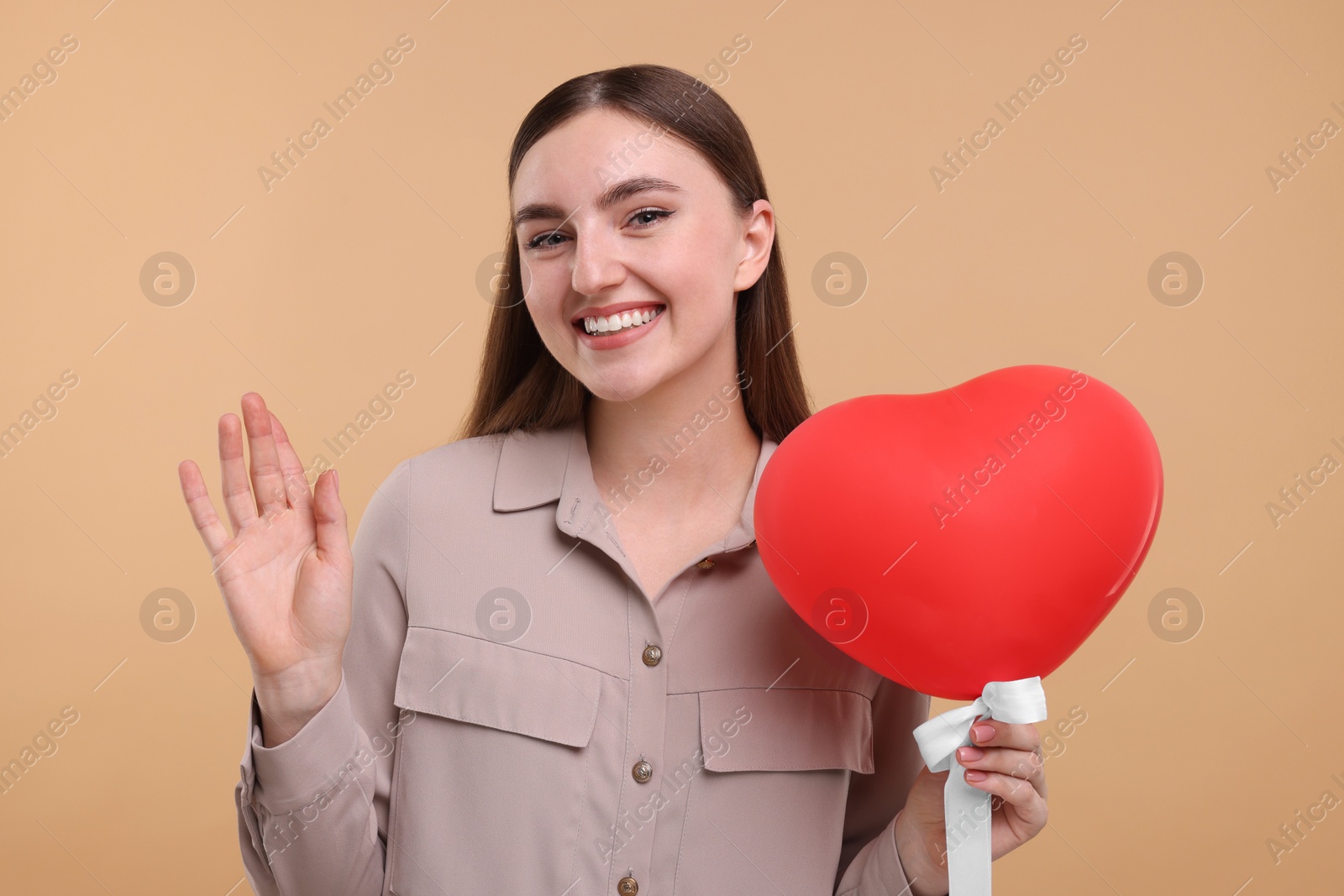 Photo of Smiling woman holding red heart shaped balloon and waving on beige background