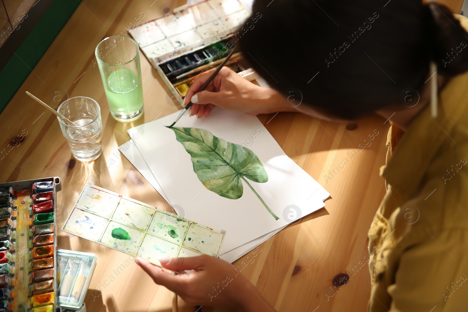 Photo of Young woman drawing leaf with watercolors at table, closeup