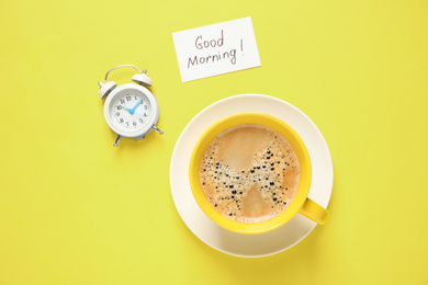 Delicious coffee, alarm clock and card with GOOD MORNING wish on yellow background, flat lay