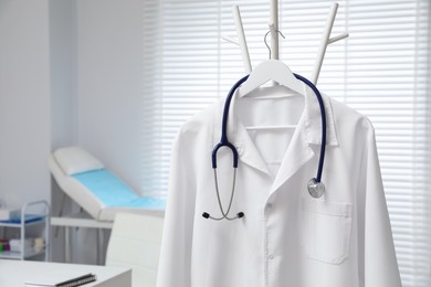 Photo of White doctor's gown and stethoscope hanging on rack in clinic, closeup. Space for text