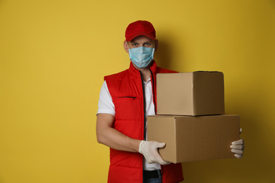 Photo of Courier in protective mask and gloves holding cardboard boxes on yellow background. Delivery service during coronavirus quarantine