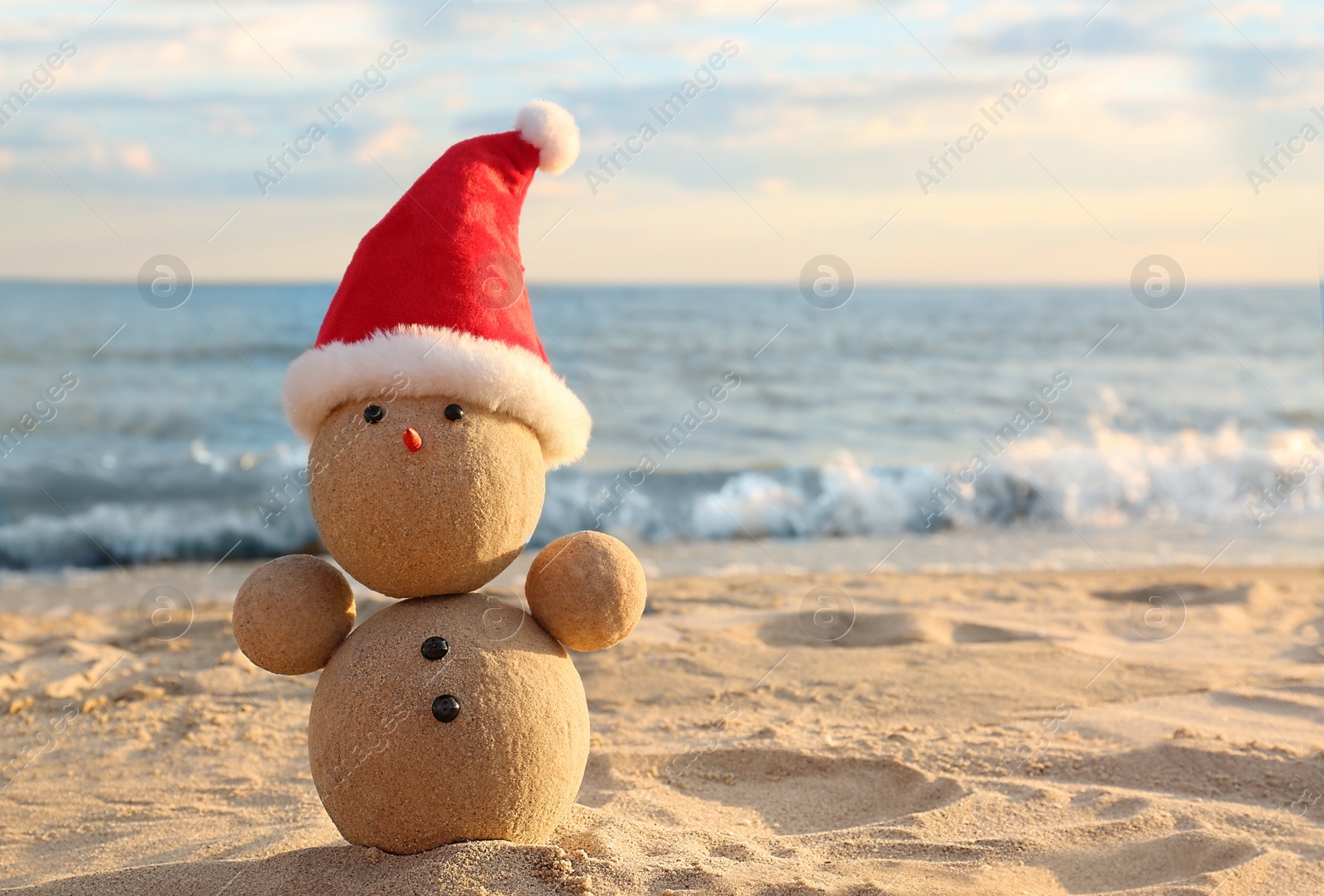 Photo of Snowman made of sand with Santa hat on beach near sea, space for text. Christmas vacation