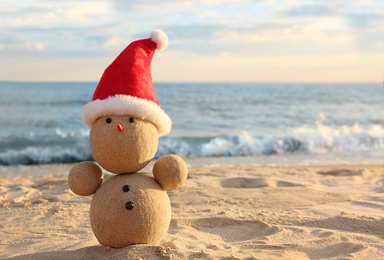 Photo of Snowman made of sand with Santa hat on beach near sea, space for text. Christmas vacation