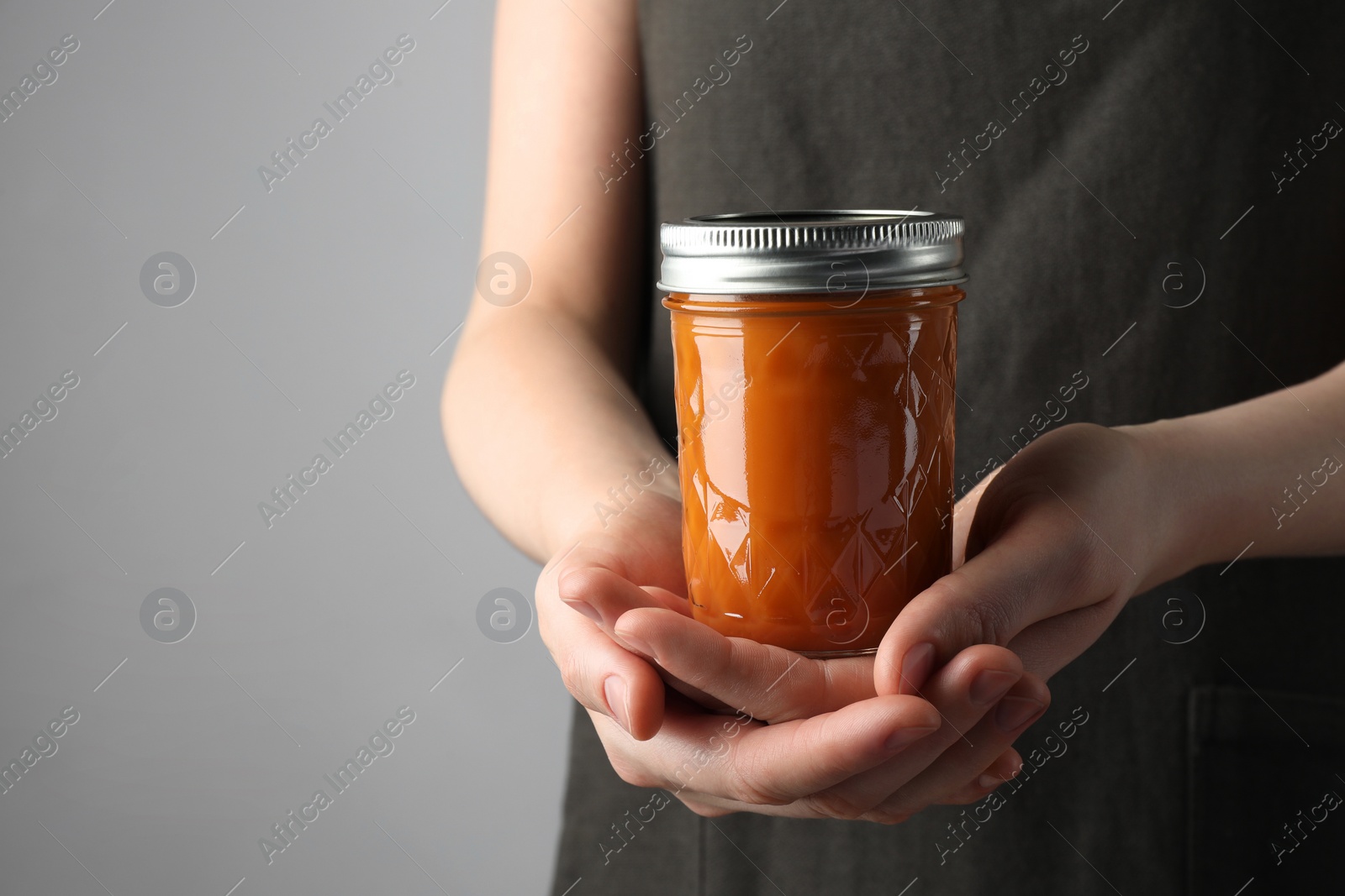 Photo of Woman holding glass jar of delicious persimmon jam on gray background, closeup. Space for text