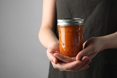 Woman holding glass jar of delicious persimmon jam on gray background, closeup. Space for text