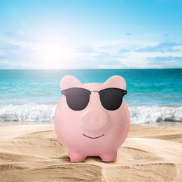 Vacation savings. Piggy bank with sunglasses on sandy beach near sea