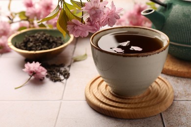 Photo of Traditional ceremony. Sakura petals in cup of brewed tea, teapot and flowers on tiled table, closeup