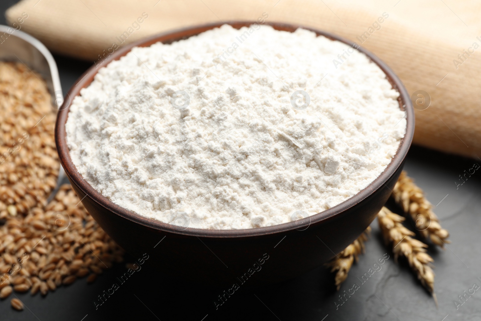 Photo of Wheat flour in bowl, spikes and grains on black table, closeup