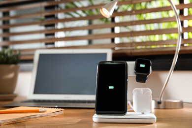 Photo of Mobile phone, earphones and smartwatch charging with wireless pad on wooden desk, space for text. Modern workplace device