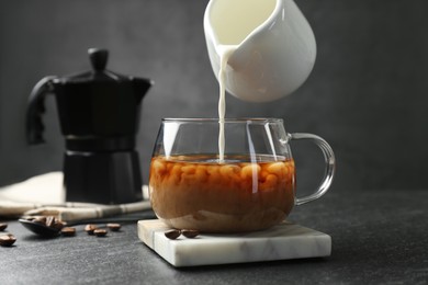 Pouring milk from pitcher into glass cup with coffee at dark textured table, closeup