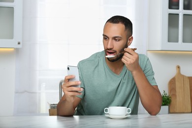 Young man using smartphone while having breakfast at white marble table in kitchen. Internet addiction