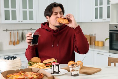 Photo of Overweight man eating tasty croissant at table in kitchen