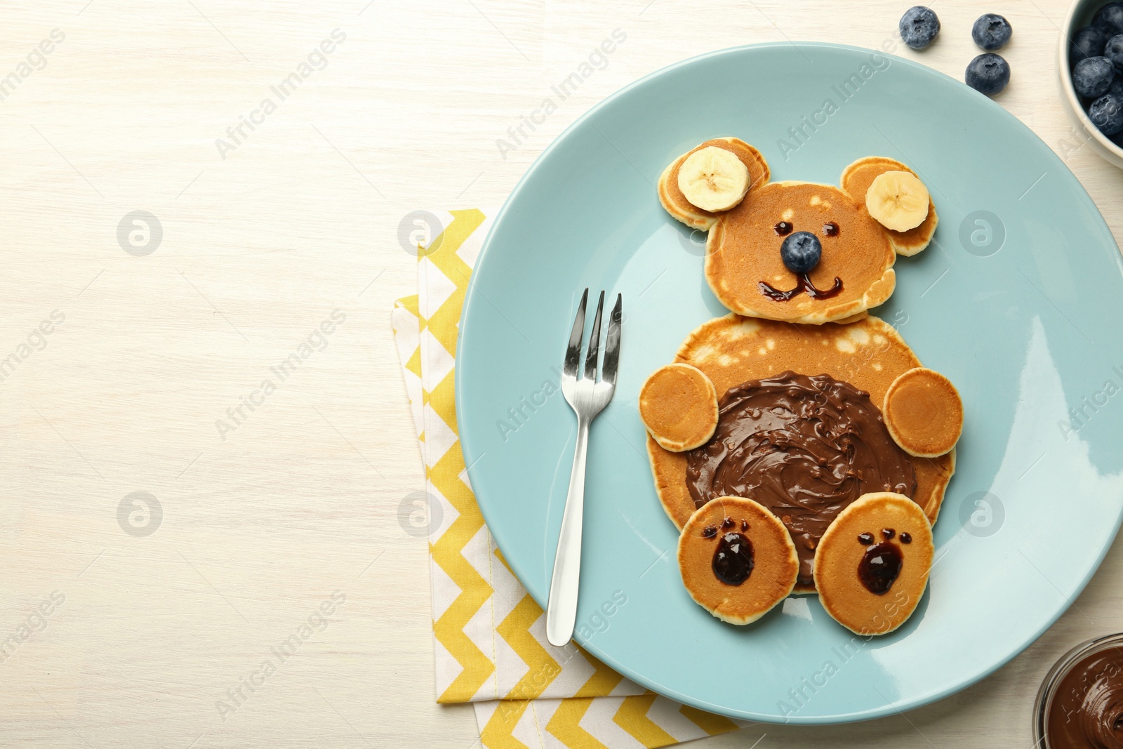 Photo of Creative serving for kids. Plate with cute bear made of pancakes, blueberries, bananas and chocolate paste on light wooden table, top view. Space for text