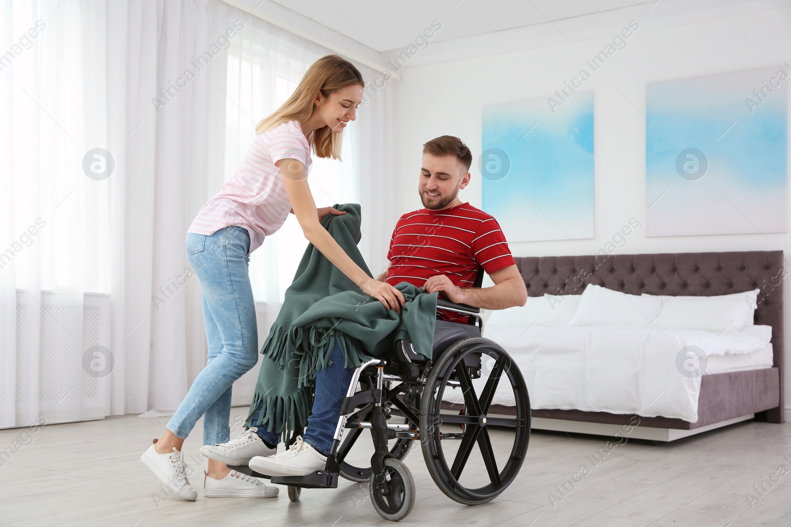 Photo of Woman covering man in wheelchair with plaid at home. Helping disabled