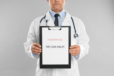 Doctor holding clipboard with words HEMORRHOID WE CAN HELP on light grey background, closeup