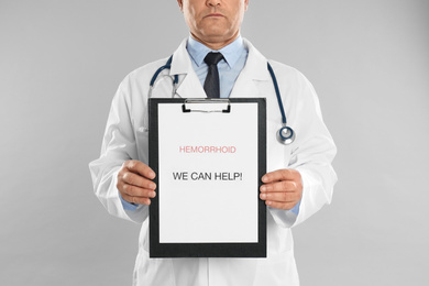 Photo of Doctor holding clipboard with words HEMORRHOID WE CAN HELP on light grey background, closeup