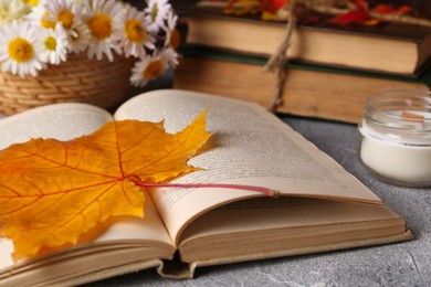 Photo of Book with autumn leaf as bookmark and scented candle on light gray textured table, closeup