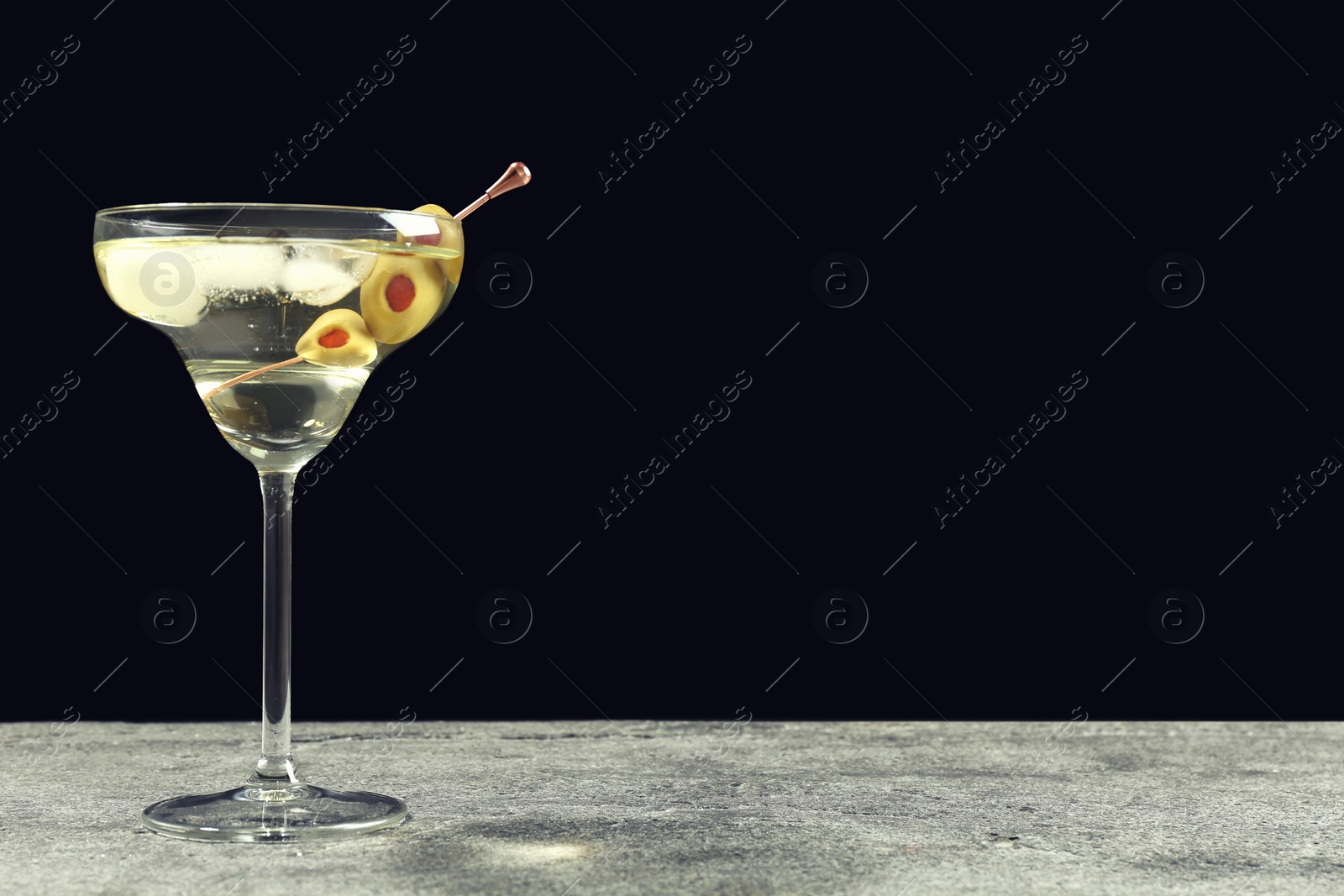 Photo of Martini cocktail with ice and olives on grey table against dark background. Space for text