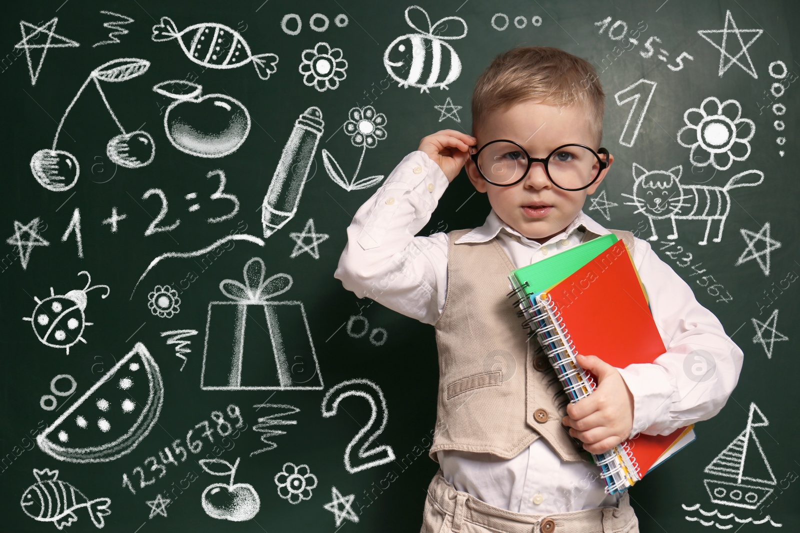 Image of Cute little child in glasses near chalkboard with different drawings. First time at school