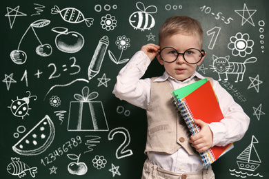 Cute little child in glasses near chalkboard with different drawings. First time at school